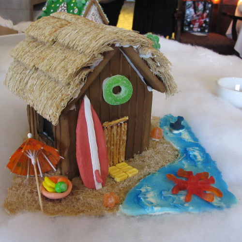 Gingerbread beach hut with surfboard, umbrella, and candy decorations on a white surface.