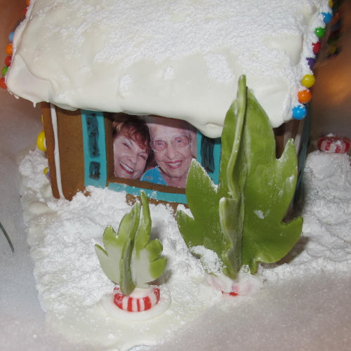 Gingerbread house with a picture of two people inside, surrounded by snow and greenery.