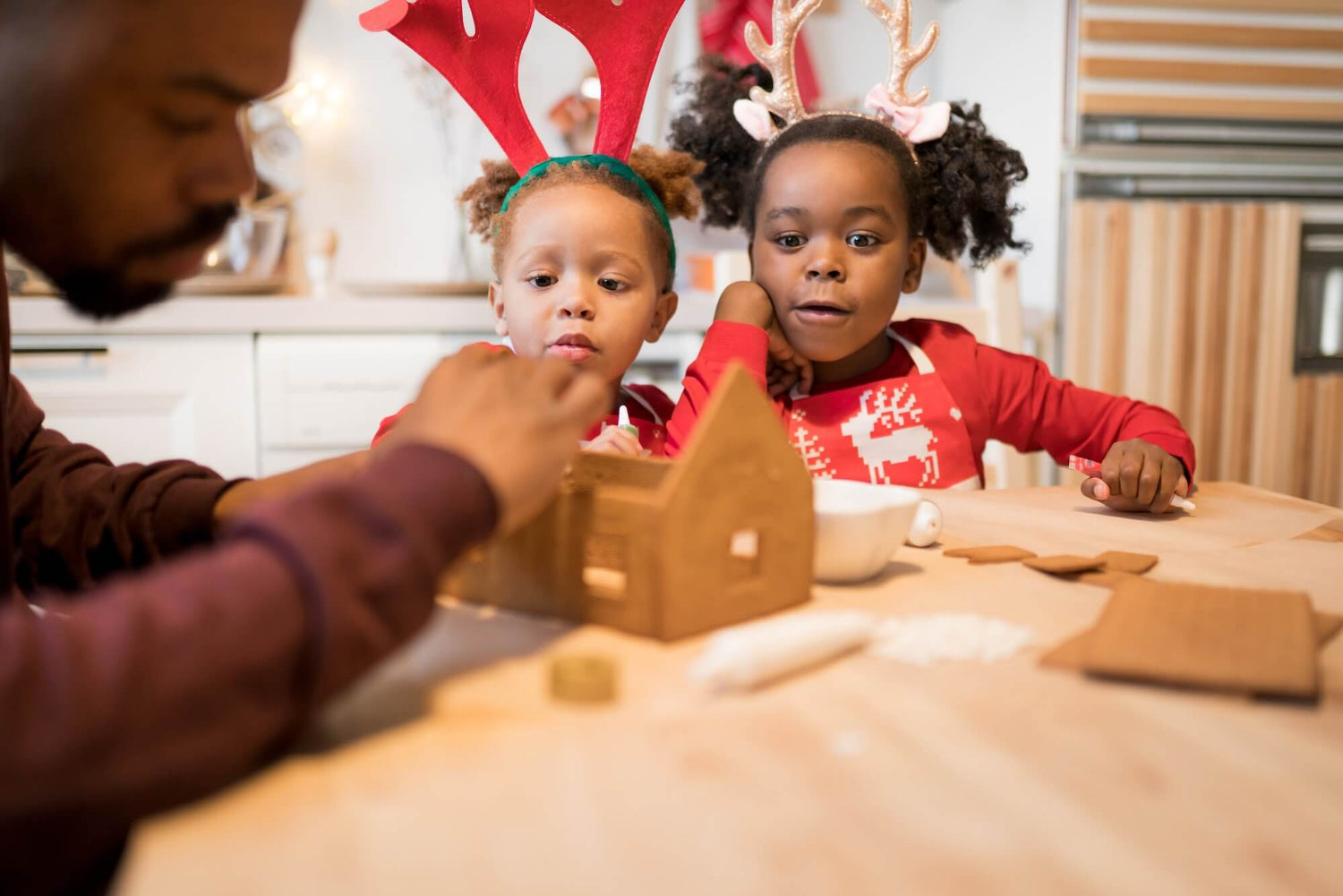 Two children and an adult sitting at a table with a gingerbread house and decorations during a holiday season.