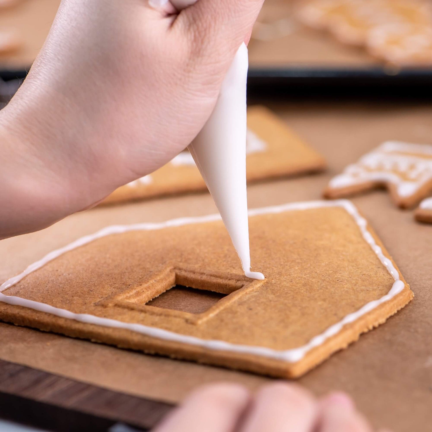 Hand decorating a gingerbread house with white icing on a wooden surface.